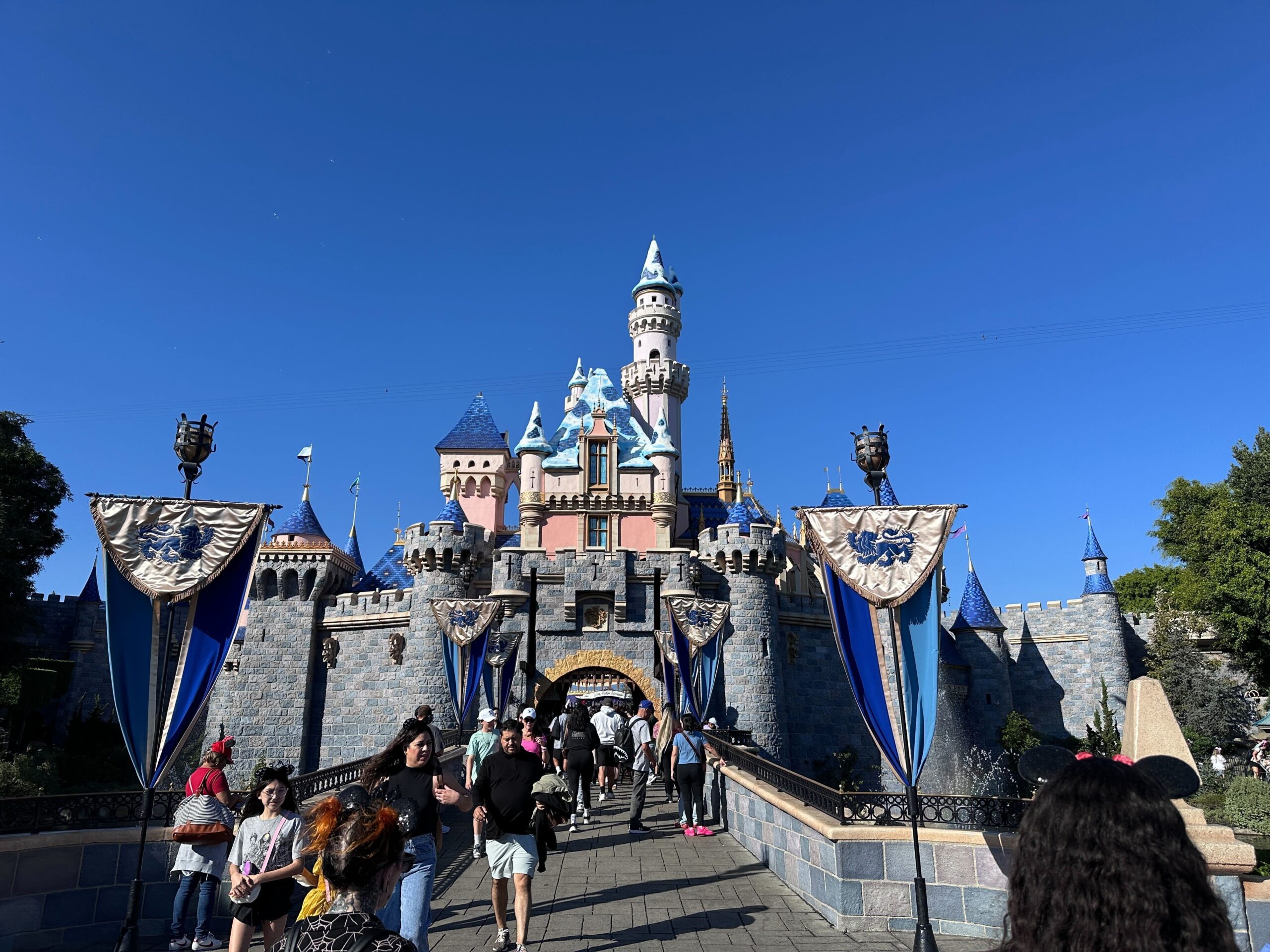 Under a clear blue sky, people walk towards the enchanting Sleeping Beauty Castle, its blue spires and intricate stonework capturing the imagination.